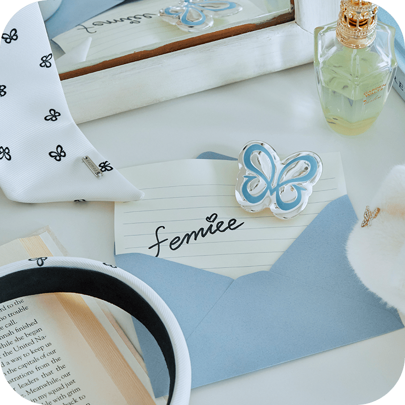 「femiee」と書かれたカードと青い封筒、蝶モチーフのヘアクリップや香水・ヘアバンドを並べた小物のフラットレイ